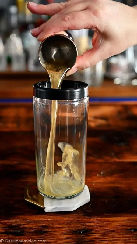 honey simple syrup being poured into a glass cocktail shaker from a jigger