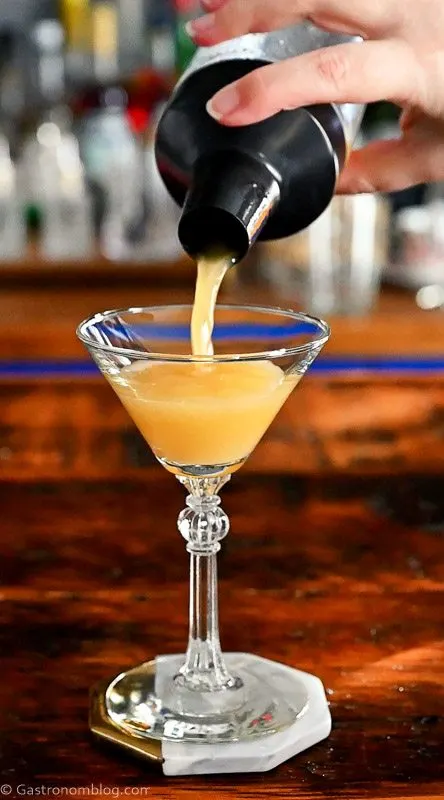 pineapple whiskey cocktail in cocktail coupe. Gold colored cocktail poured from cocktail shaker.