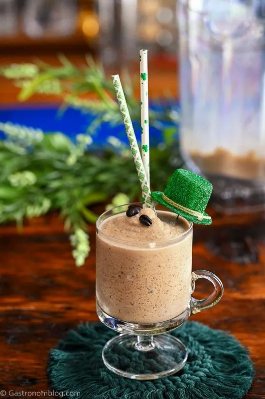tan cocktail in footed glass mug, this Frozen Irish Coffee has St Patrick's Day straws, hat and greenery behind