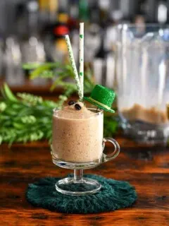 tan cocktail in footed glass mug, this Frozen Irish Coffee has St Patrick's Day straws, hat and greenery behind