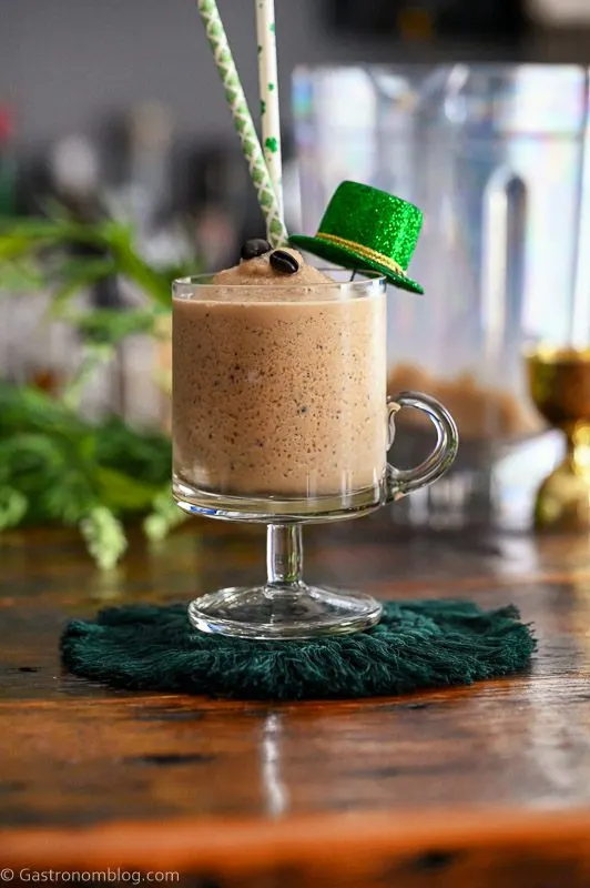 tan cocktail in footed glass mug, this Frozen Irish Coffee has St Patrick's Day straws, hat and greenery behind