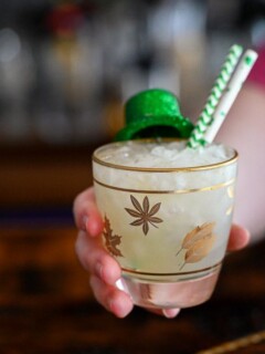 hand holding Irish Mule cocktail in gold leaf rocks glass with St Patricks Day clover straws and hat garnish, gold barware behind