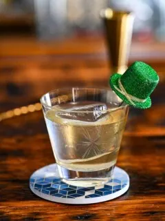 gold colored Irish old fashioned cocktail in rocks glass with green glitter hat garnish on blue coaster on wooden table