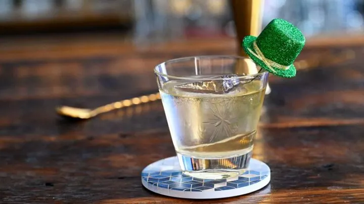 gold colored Irish old fashioned cocktail in rocks glass with green glitter hat garnish on blue coaster on wooden table