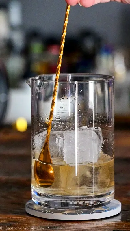 gold bar spoon stirring ice in mixing glass with tan Old Fashioned cocktail