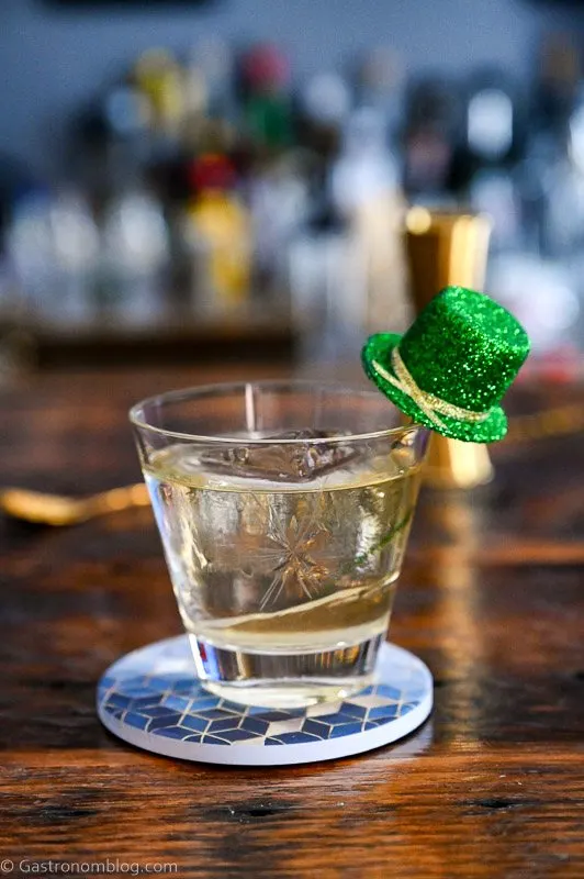 gold colored Irish old fashioned cocktail in rocks glass with green glitter hat garnish on blue coaster on wooden table