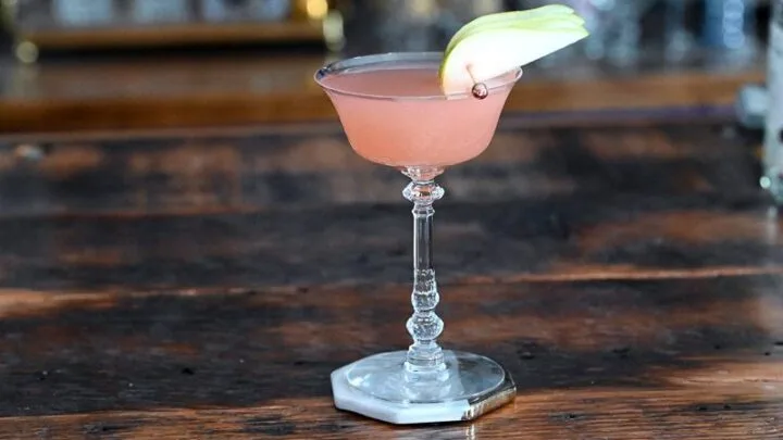 pink Grapefruit Gin Cocktail in a glass cocktail coupe, pear slice garnish on a white and gold coaster on a wood table
