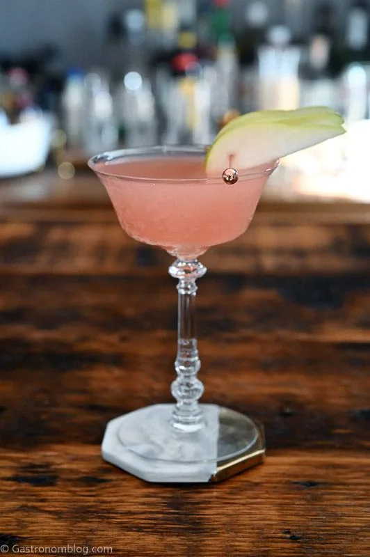 pink Grapefruit Gin Cocktail in a glass cocktail coupe, pear slice garnish on a white and gold coaster on a wood table
