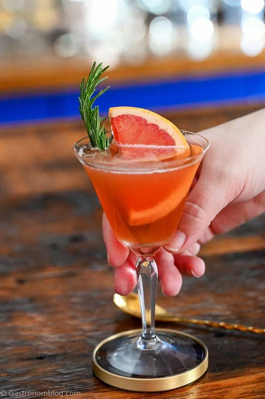 Orange colored cocktail in coupe glass. Bourbon Grapefruit cocktail garnished with rosemary and grapefruit slice on wooden table.
