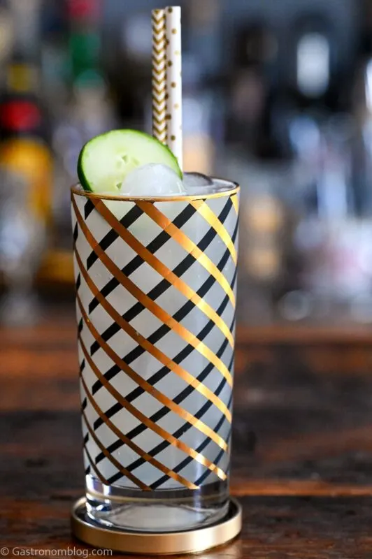 opaque Cucumber Collins in gold and black striped highball glass, cucumber slice and gold straw on top. On wood table