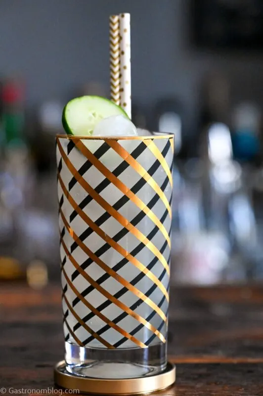 opaque Cucumber Collins in gold and black striped highball glass, cucumber slice and gold straw on top. On wood table