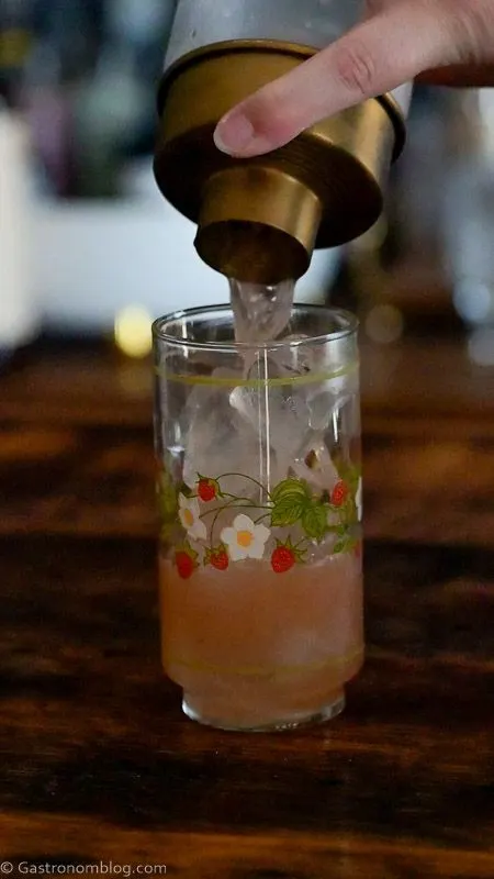 pink strawberry vanilla cocktail being poured into strawberry printed glass from a gold and glass cocktail shaker