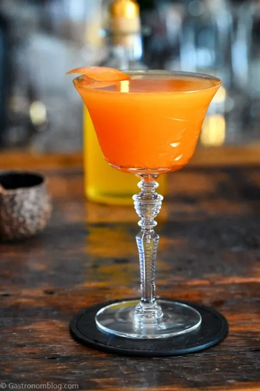 orange colored cocktail in a tall clear coupe glass on a wooden table, bar tools behind