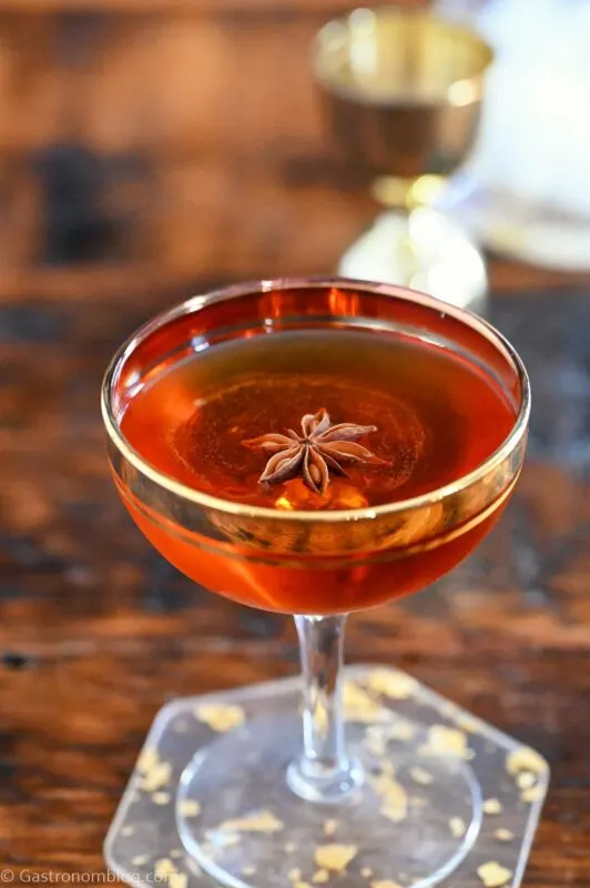 Red colored Fall Apple Cocktail in gold rimmed coupe glass on gold speckled clear coaster on wood table, star anise garnish