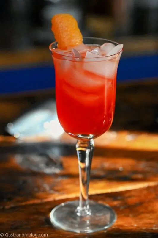 Pink colored cocktail in tall wine glass with ice and orange peel on wood table.