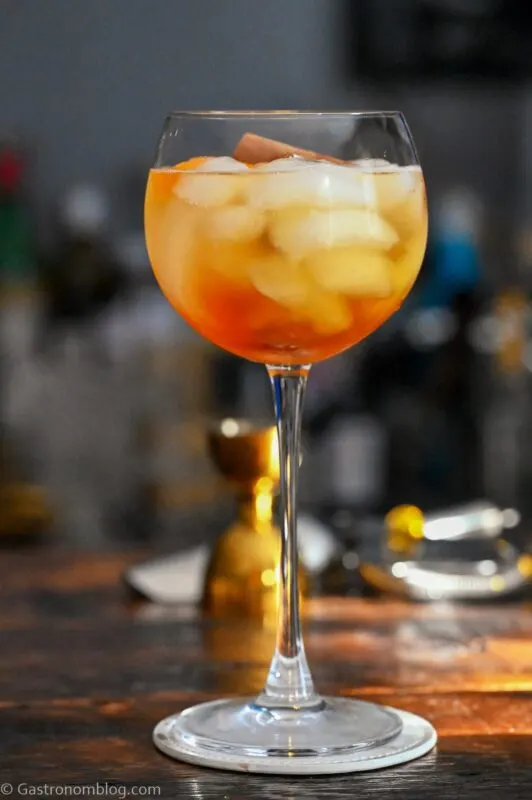 SPRITZ Musim Dingin Berwarna Emas dalam gelas anggur tinggi dengan es batu. Pada coaster putih di atas meja kayu.