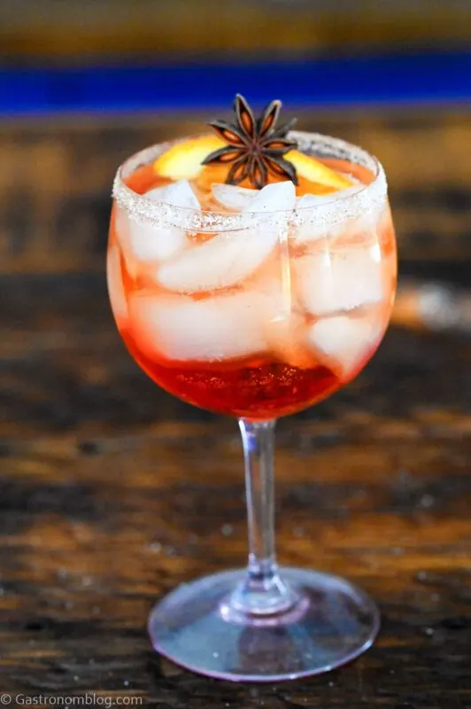 red Fall Aperol Spritz in wine glass with ice cubes, star anise and orange garnish. On wood table.