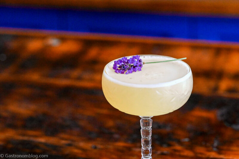 Yellow cocktail in clear cocktail coupe. This Lavender Bees Knees has a lavender sprig garnish, is on a coaster on a wooden table.