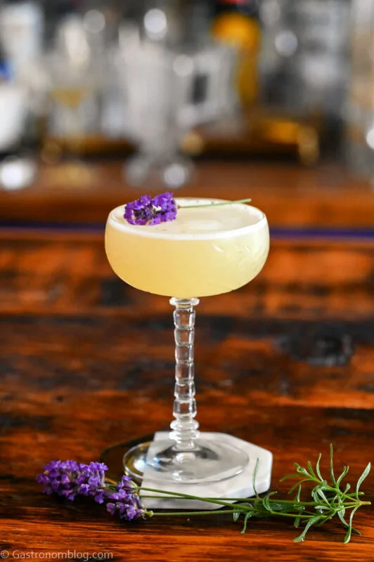 Yellow cocktail in clear cocktail coupe. This Lavender Bees Knees has a lavender sprig garnish, is on a coaster on a wooden table.