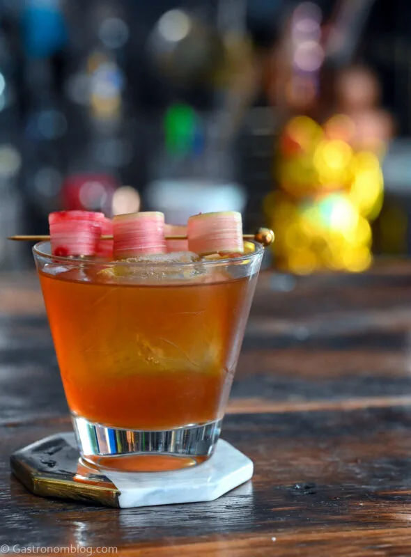 brown cocktail in rocks glass with rhubarb slice garnish on pick. Glass is on a coaster on a wooden table.