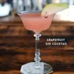 pink Grapefruit Gin Cocktail in a glass cocktail coupe, pear slice garnish on a white and gold coaster on a wood table