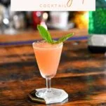 pink colored lavender syrup cocktail in a tall coupe with a basil leaf garnish on white coaster on wood table.