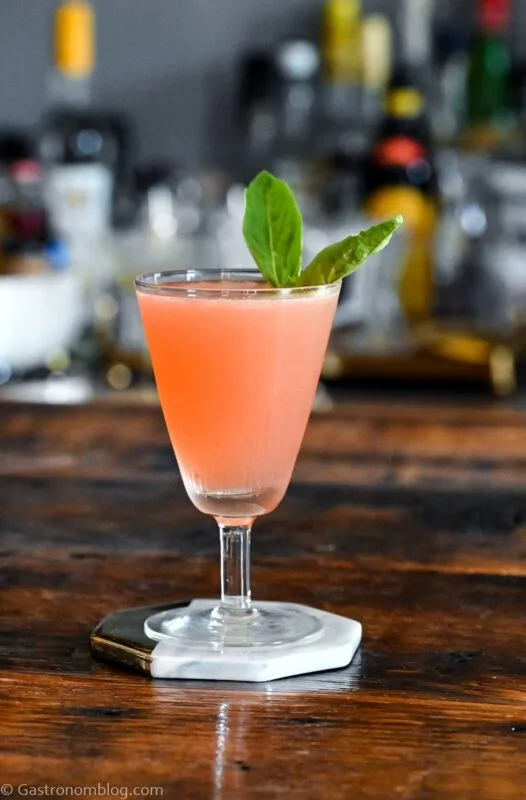 pink colored lavender syrup cocktail in a tall coupe with a basil leaf garnish on white coaster on wood table.