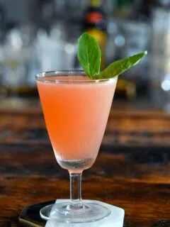 pink colored lavender syrup cocktail in a tall coupe with a basil leaf garnish on white coaster on wood table.