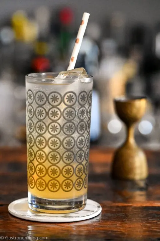 orange and white colored Earl Grey Collins cocktail in gray circle printed highball glass, on white coaster on wooden table