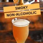 Orange colored Smoky mocktail in tall cocktail coupe with foam on white and gold coaster on wooden table