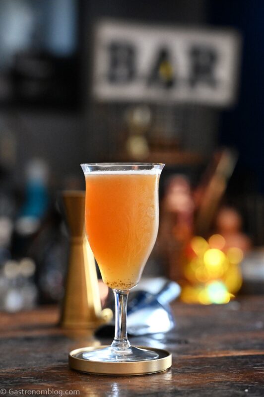 Orange colored Smoky mocktail in tall cocktail coupe with foam on white and gold coaster on wooden table