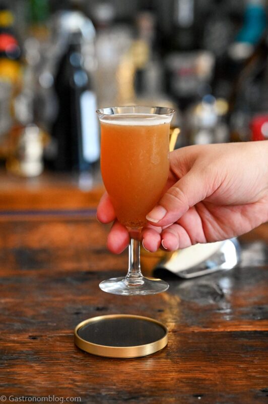Orange colored Smoky mocktail in tall cocktail coupe with foam on white and gold coaster on wooden table