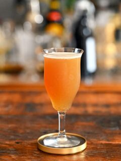 Orange colored Smoky mocktail in tall cocktail coupe with foam on white and gold coaster on wooden table