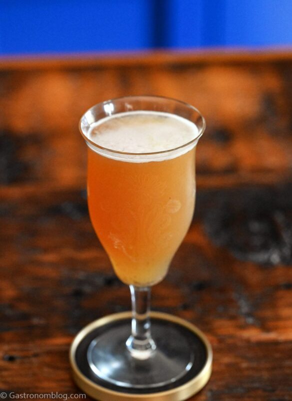 Orange colored Smoky mocktail in tall cocktail coupe with foam on white and gold coaster on wooden table
