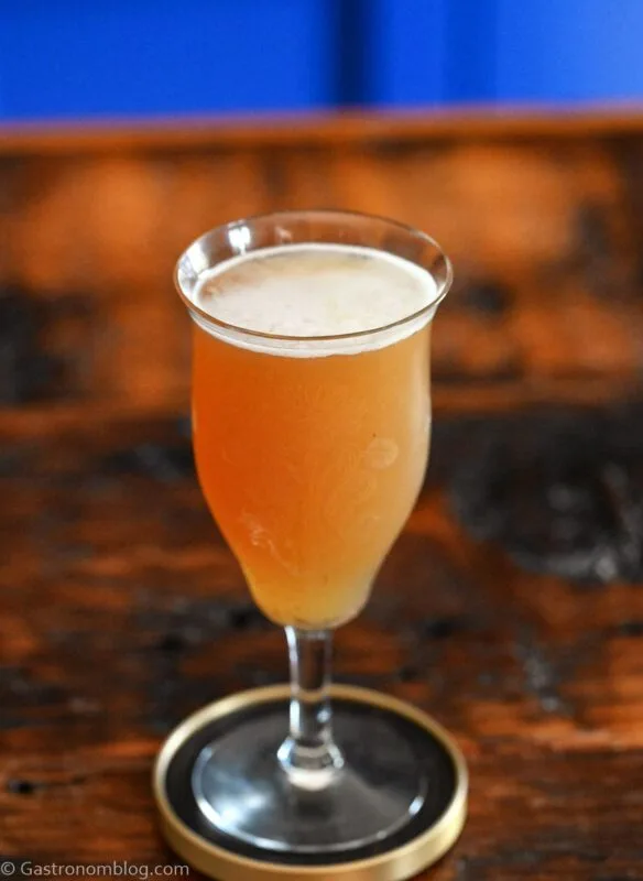 Orange colored Smoky mocktail in tall cocktail coupe with foam on white and gold coaster on wooden table