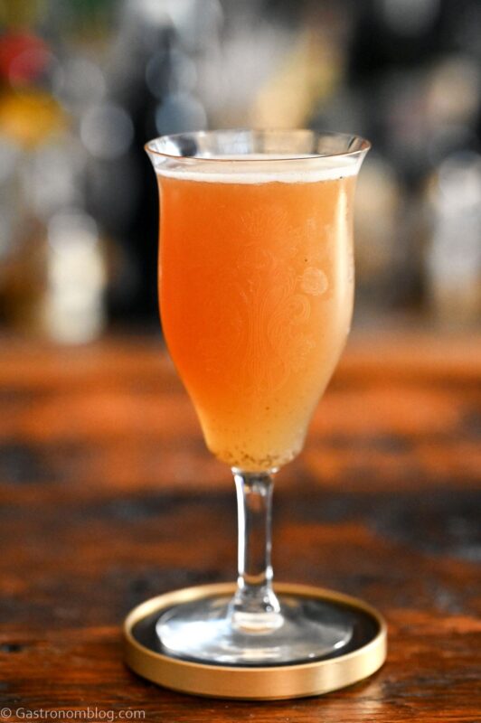 Orange colored Smoky mocktail in tall cocktail coupe with foam on white and gold coaster on wooden table
