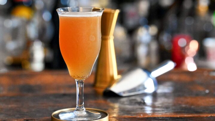 Orange colored Smoky mocktail in tall cocktail coupe with foam on white and gold coaster on wooden table