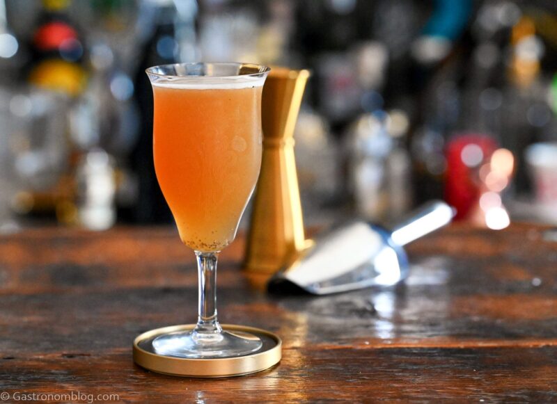 Orange colored Smoky mocktail in tall cocktail coupe with foam on white and gold coaster on wooden table