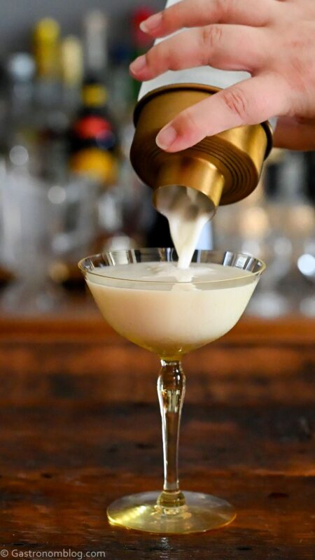pouring white yogurt cocktail from cocktail shaker in to yellow cocktail coupe