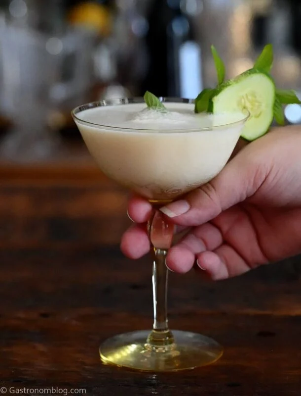 hand holding yellow cocktail glass with white yogurt cocktail and cucumber and mint garnish