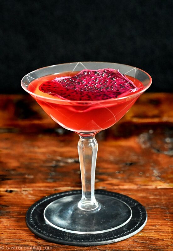 red Stranger Things Drink with dried dragonfruit chip garnish in cocktail coupe on wooden table with black background